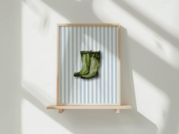 A framed art print featuring a pair of green rain boots on a light blue and white striped background, displayed on a light wood floating shelf against a white wall with soft window shadows.