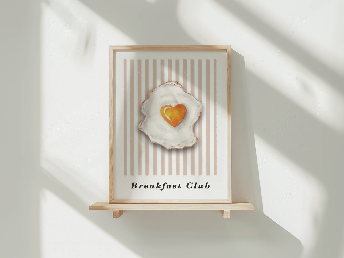 A framed art print featuring a stylized fried egg with a heart-shaped yolk, displayed on a light wooden stand against a white wall with soft window shadows.