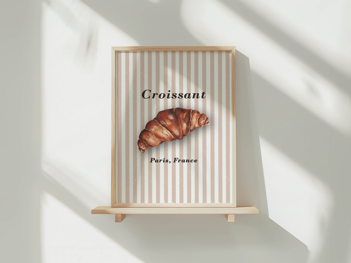 A framed wall art print featuring a croissant illustration with the text 'Croissant' and 'Paris, France' on a beige and white striped background, displayed on a light wood shelf against a white wall with soft window shadows.