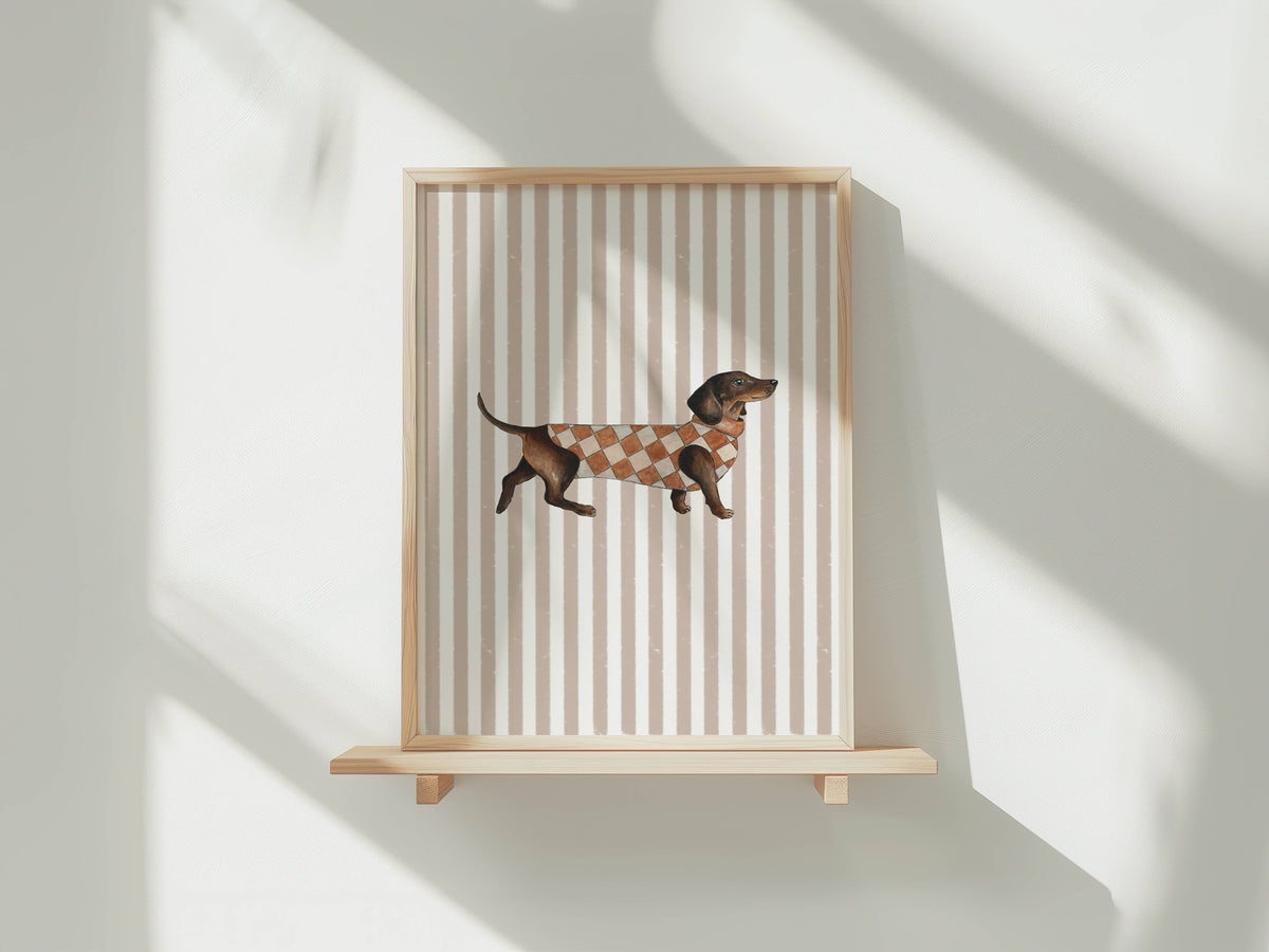 A framed art print featuring a dachshund wearing a brown and beige argyle sweater, displayed on a light wood floating shelf against a white wall with soft diagonal shadows.