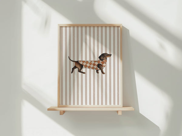 A framed art print featuring a dachshund wearing a brown and beige argyle sweater, displayed on a light wood floating shelf against a white wall with soft diagonal shadows.