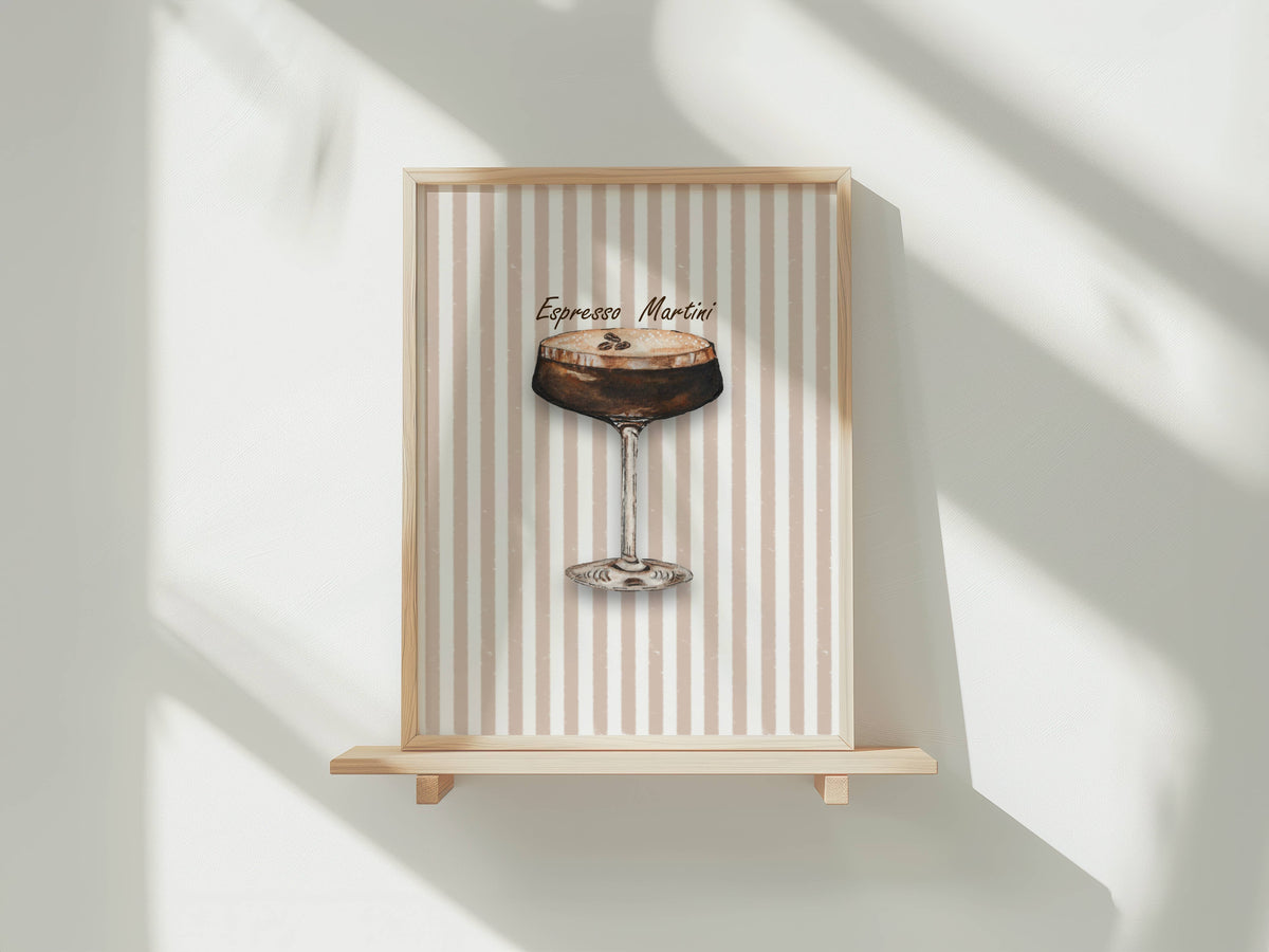 A framed art print featuring an espresso martini cocktail illustration on a beige and white striped background, displayed on a light wood floating shelf against a white wall with soft window shadows.