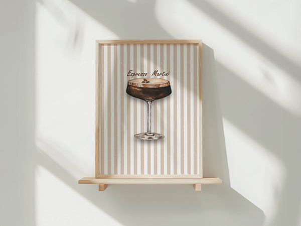 A framed art print featuring an espresso martini cocktail illustration on a beige and white striped background, displayed on a light wood floating shelf against a white wall with soft window shadows.