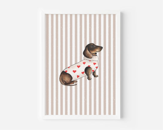 A framed art print featuring a watercolor illustration of a dachshund wearing a white sweater with red hearts, set against a background of vertical beige and white stripes.