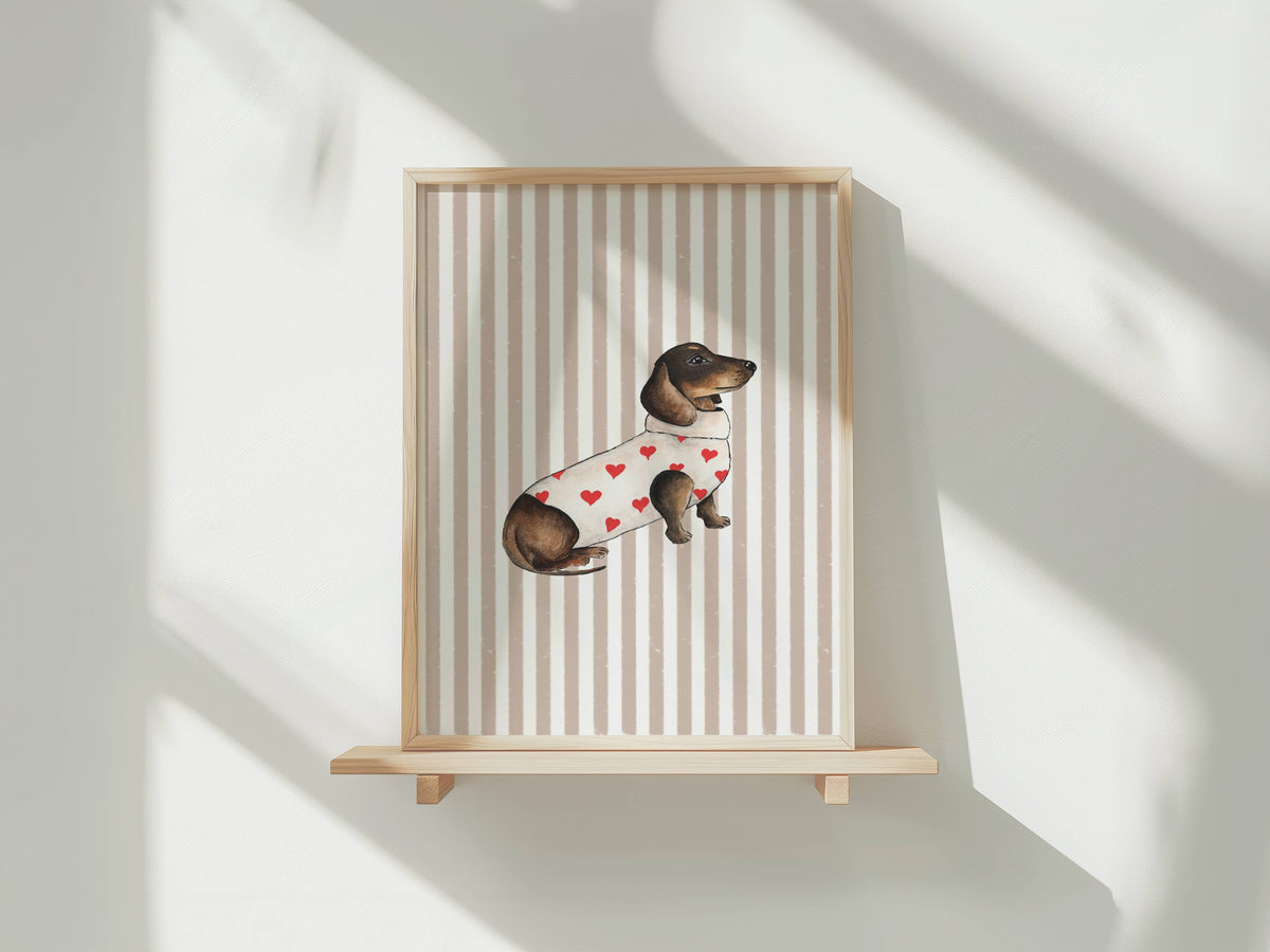 A framed art print featuring a watercolor illustration of a dachshund wearing a white sweater with red hearts, displayed on a light wood shelf against a white wall with soft window shadows.