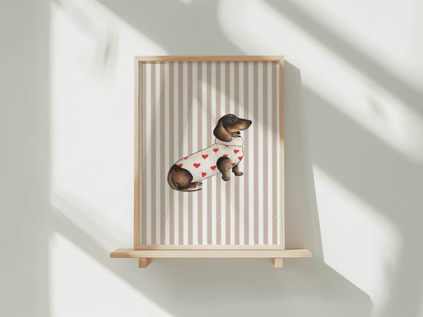 A framed art print featuring a watercolor illustration of a dachshund wearing a white sweater with red hearts, displayed on a light wood shelf against a white wall with soft window shadows.