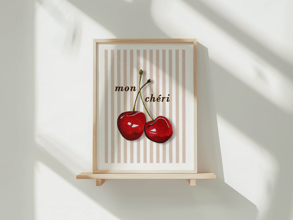 A framed art print featuring two red cherries on a cream and beige striped background with the French text 'mon chéri' above them, displayed on a light wooden shelf against a white wall with soft window shadows.