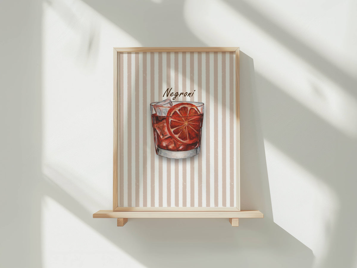 A framed art print featuring a watercolour illustration of a Negroni cocktail in a tumbler glass with ice and an orange slice garnish, set against a background of vertical beige and white stripes. The print is shown in a light wood frame displayed on a wooden shelf against a white wall with soft window shadows.