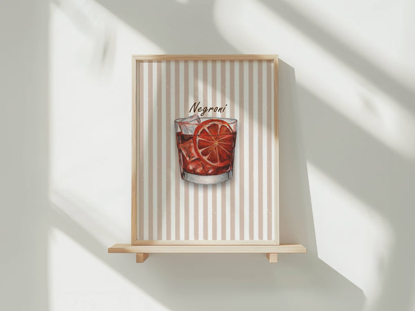 A framed art print featuring a watercolour illustration of a Negroni cocktail in a tumbler glass with ice and an orange slice garnish, set against a background of vertical beige and white stripes. The print is shown in a light wood frame displayed on a wooden shelf against a white wall with soft window shadows.