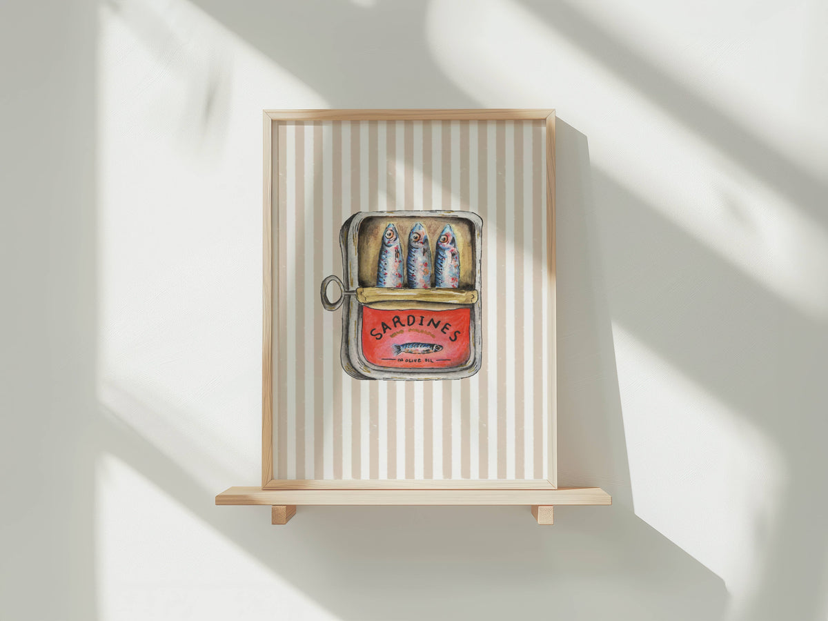 A framed art print featuring a vintage-style illustration of three sardines in a red can labeled 'SARDINES', mounted on a light wood shelf against a white wall with soft diagonal shadows.