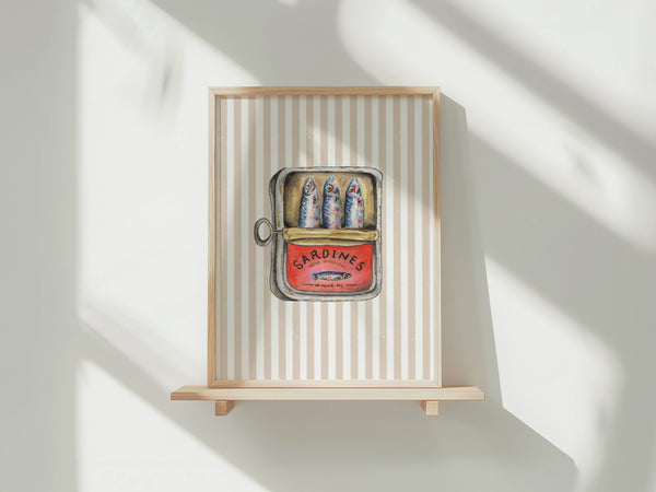 A framed art print featuring a vintage-style illustration of three sardines in a red can labeled 'SARDINES', mounted on a light wood shelf against a white wall with soft diagonal shadows.