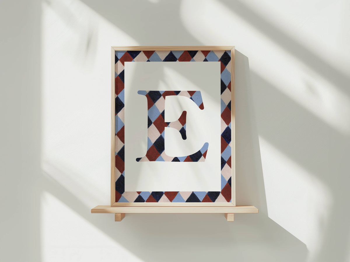 A framed letter art print featuring a stylized capital letter E with a geometric diamond pattern in navy, burgundy, and light blue, mounted on a light wood frame and displayed on a wooden shelf against a white wall with soft window shadows.