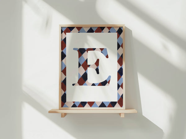 A framed letter art print featuring a stylized capital letter E with a geometric diamond pattern in navy, burgundy, and light blue, mounted on a light wood frame and displayed on a wooden shelf against a white wall with soft window shadows.