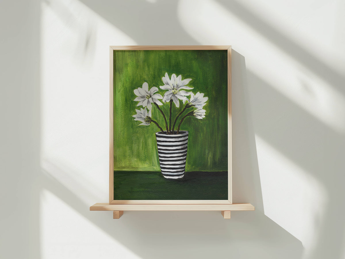 A framed painting of white flowers in a black-and-white striped vase against a green background, displayed on a light wood shelf against a white wall with soft window shadows.