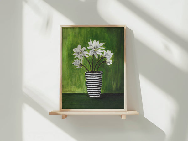 A framed painting of white flowers in a black-and-white striped vase against a green background, displayed on a light wood shelf against a white wall with soft window shadows.