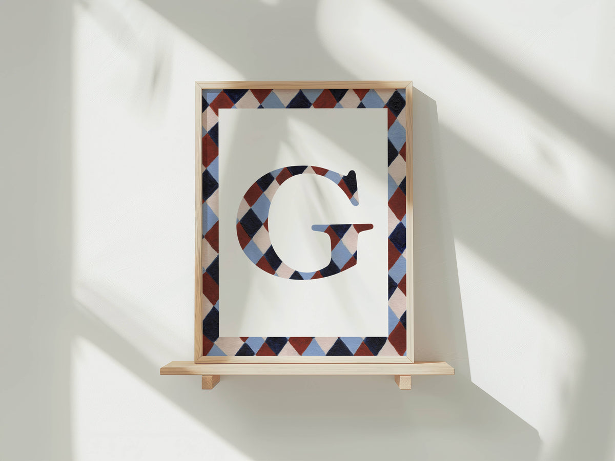 A framed art print featuring a large decorative letter 'G' in a geometric diamond pattern, displayed on a light wooden shelf against a white wall with soft window shadows.
