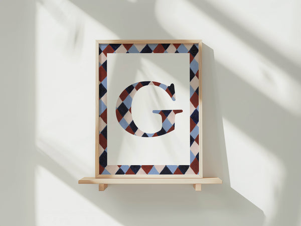 A framed art print featuring a large decorative letter 'G' in a geometric diamond pattern, displayed on a light wooden shelf against a white wall with soft window shadows.