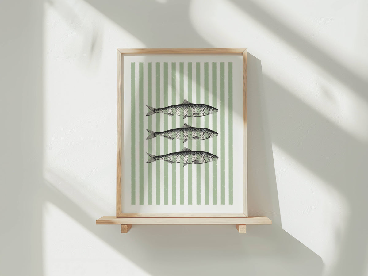 Framed wall art featuring three fish illustrations on a white background with vertical sage green stripes, displayed on a light wood shelf against a white wall with soft window shadows.