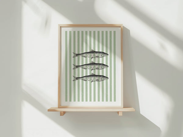 Framed wall art featuring three fish illustrations on a white background with vertical sage green stripes, displayed on a light wood shelf against a white wall with soft window shadows.