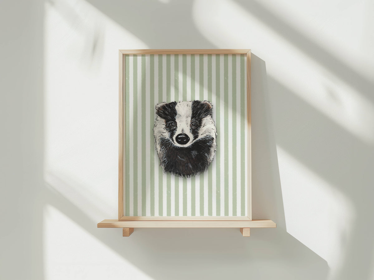 A framed art print featuring a black and white badger portrait on a light green and white striped background, displayed on a light wood floating shelf against a white wall with soft window shadows.