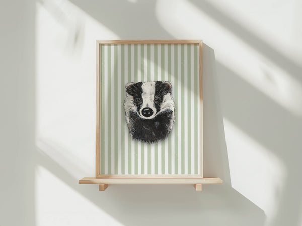 A framed art print featuring a black and white badger portrait on a light green and white striped background, displayed on a light wood floating shelf against a white wall with soft window shadows.