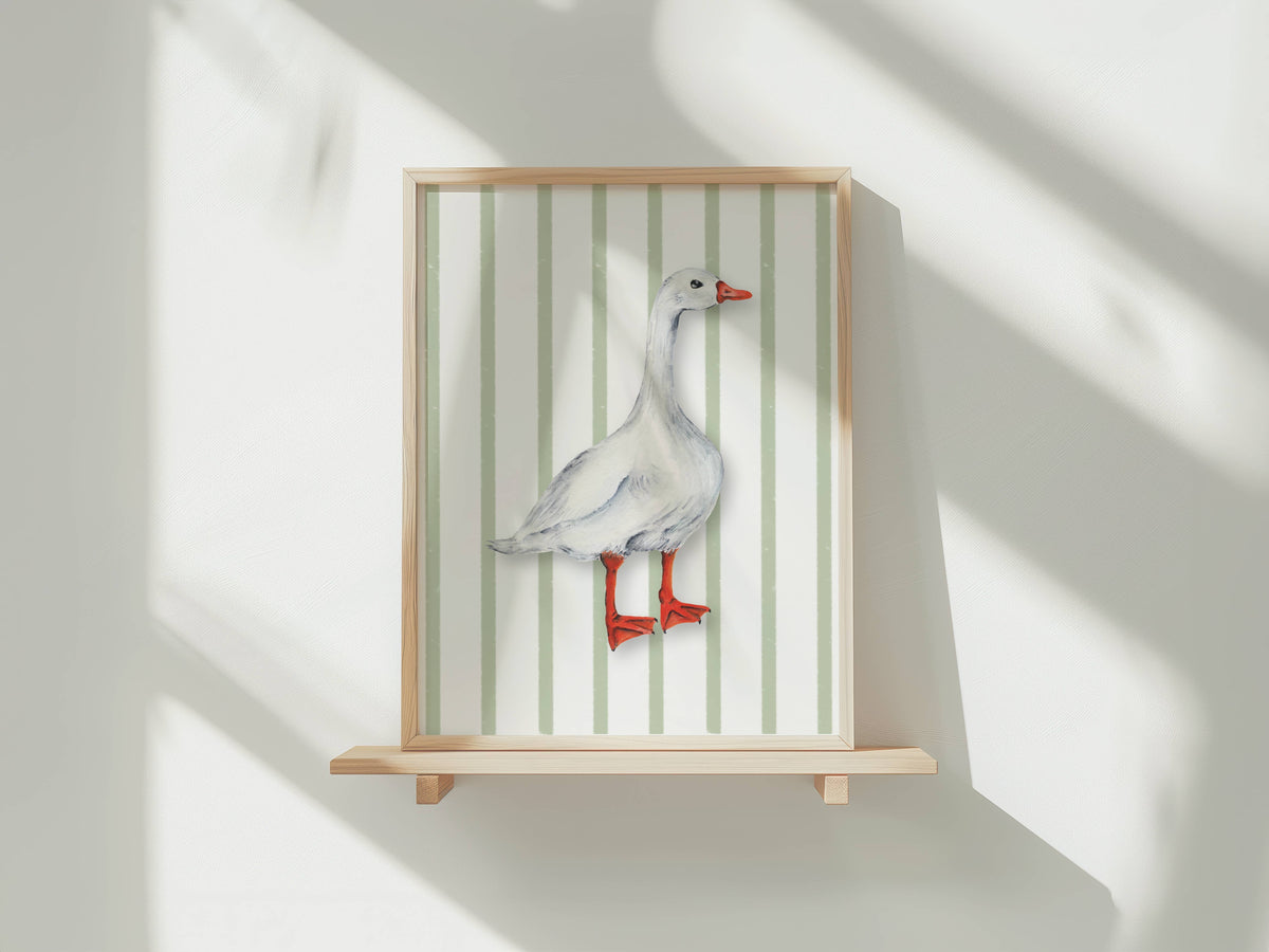 A framed art print featuring a watercolour illustration of a white goose with an orange-red beak and webbed feet, set against a background of vertical pale green and white stripes. The print is shown in a light wood frame displayed on a wooden shelf against a white wall with soft window shadows.