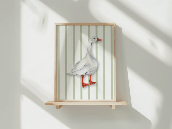 A framed art print featuring a watercolour illustration of a white goose with an orange-red beak and webbed feet, set against a background of vertical pale green and white stripes. The print is shown in a light wood frame displayed on a wooden shelf against a white wall with soft window shadows.