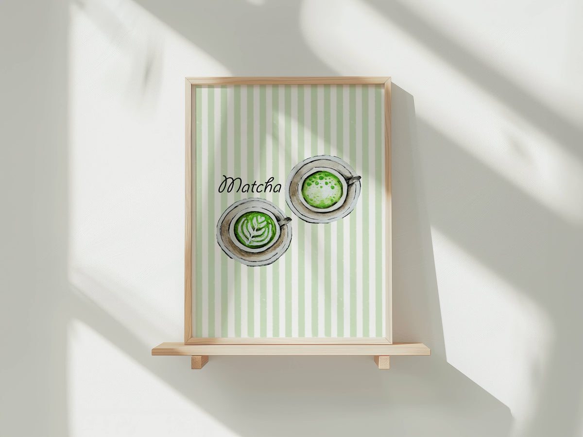 A framed art print featuring a watercolour illustration of two matcha lattes viewed from above with latte art, set against a background of vertical pale green and white stripes, with the word 'Matcha' in black script. The print is shown in a light wood frame displayed on a wooden shelf against a white wall with soft window shadows.