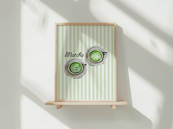 A framed art print featuring a watercolour illustration of two matcha lattes viewed from above with latte art, set against a background of vertical pale green and white stripes, with the word 'Matcha' in black script. The print is shown in a light wood frame displayed on a wooden shelf against a white wall with soft window shadows.