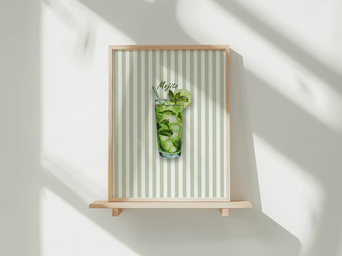 A framed art print featuring a watercolour illustration of a mojito cocktail in a tall glass with mint leaves, ice and a lime wheel, set against a background of vertical pale green and white stripes. The print is shown in a light wood frame displayed on a wooden shelf against a white wall with soft window shadows.