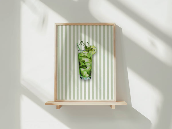 A framed art print featuring a watercolour illustration of a mojito cocktail in a tall glass with mint leaves, ice and a lime wheel, set against a background of vertical pale green and white stripes. The print is shown in a light wood frame displayed on a wooden shelf against a white wall with soft window shadows.