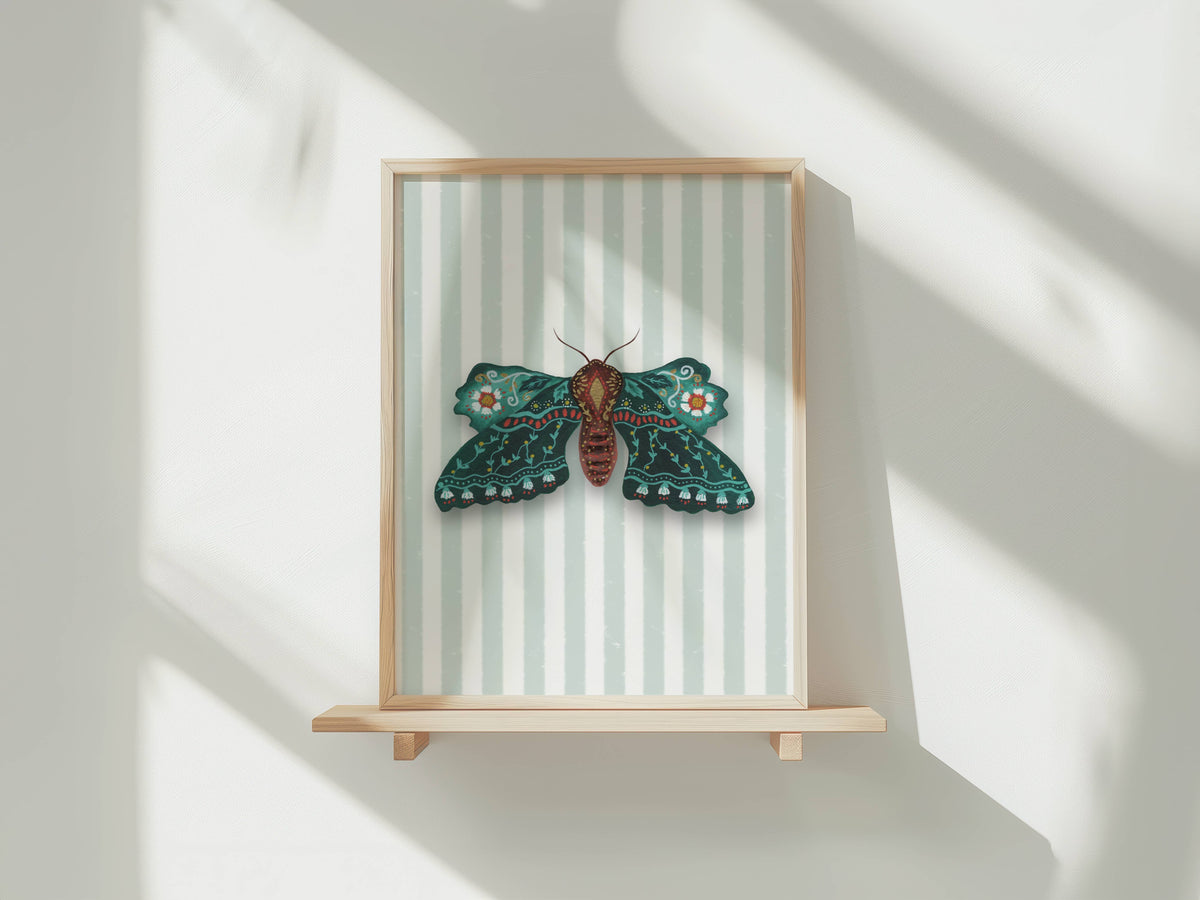A framed wall art piece featuring a detailed green and brown moth on a pale striped background, displayed on a light wood shelf against a white wall with soft window shadows.