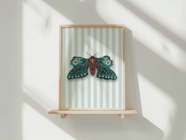 A framed wall art piece featuring a detailed green and brown moth on a pale striped background, displayed on a light wood shelf against a white wall with soft window shadows.