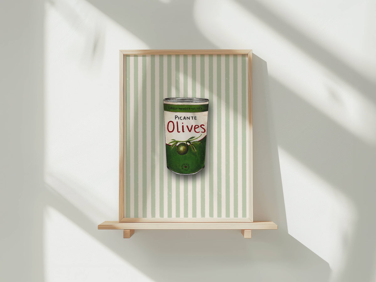 A framed art print featuring a watercolour illustration of a tin of Picante Olives with a green label, set against a background of vertical pale green and white stripes. The print is shown in a light wood frame displayed on a wooden shelf against a white wall with soft window shadows.