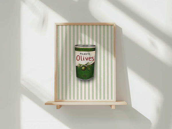 A framed art print featuring a watercolour illustration of a tin of Picante Olives with a green label, set against a background of vertical pale green and white stripes. The print is shown in a light wood frame displayed on a wooden shelf against a white wall with soft window shadows.