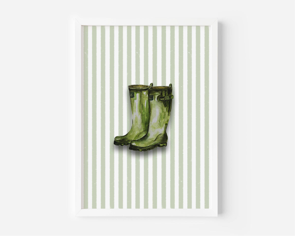A framed art print featuring a pair of green rain boots centered on a background of vertical light green and white stripes, displayed against a clean white wall.