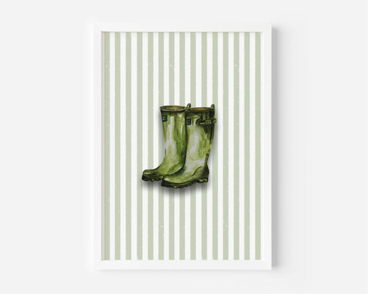 A framed art print featuring a pair of green rain boots centered on a background of vertical light green and white stripes, displayed against a clean white wall.