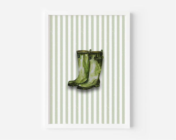A framed art print featuring a pair of green rain boots centered on a background of vertical light green and white stripes, displayed against a clean white wall.