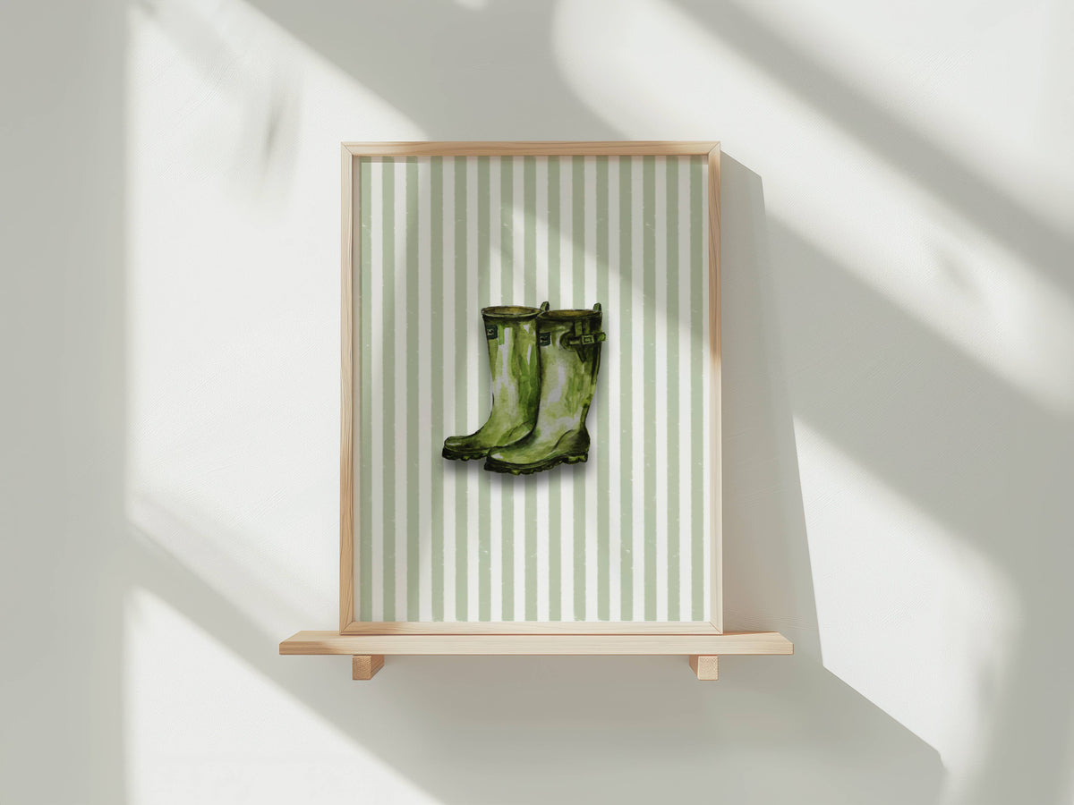A framed art print featuring a pair of green rain boots on a light green and white striped background, displayed on a light wood floating shelf against a white wall with soft window shadows.