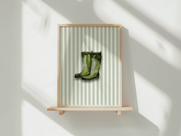 A framed art print featuring a pair of green rain boots on a light green and white striped background, displayed on a light wood floating shelf against a white wall with soft window shadows.