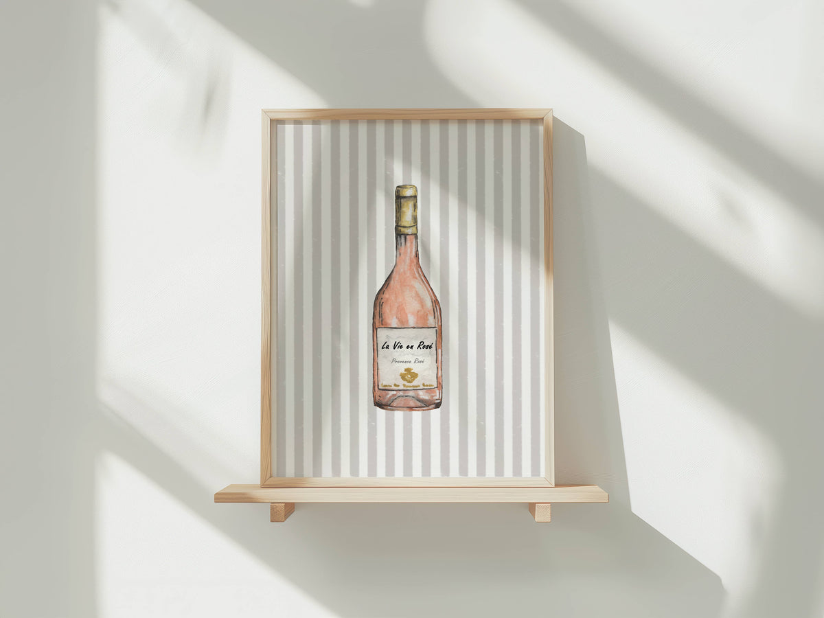 A framed art print featuring a watercolor illustration of a rosé wine bottle, displayed on a light wood floating shelf against a white wall with soft window shadows.