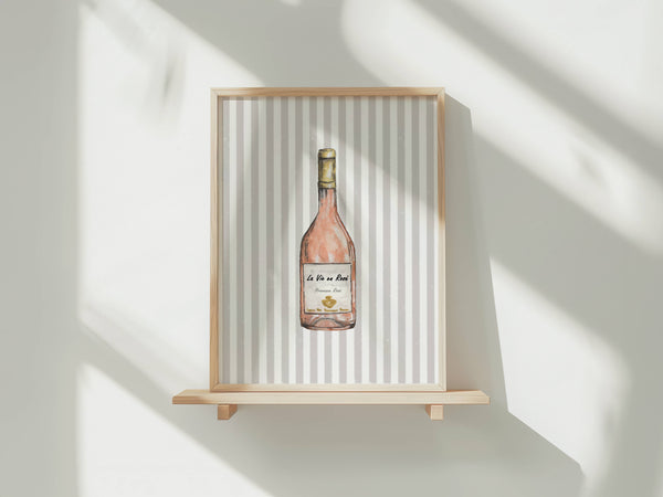 A framed art print featuring a watercolor illustration of a rosé wine bottle, displayed on a light wood floating shelf against a white wall with soft window shadows.