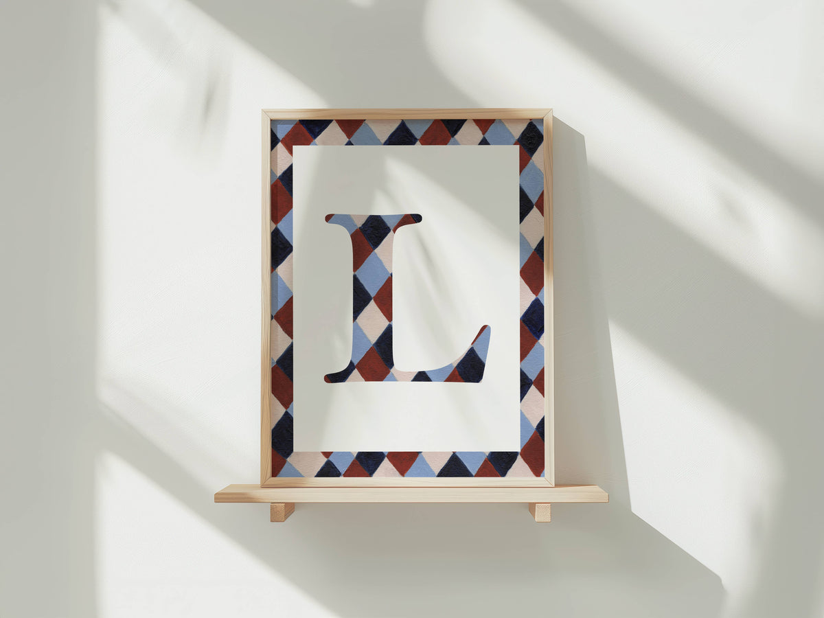 A framed letter monogram print featuring a large capital letter 'L' in a geometric diamond pattern, mounted on a light wood shelf against a white wall with soft window shadows.