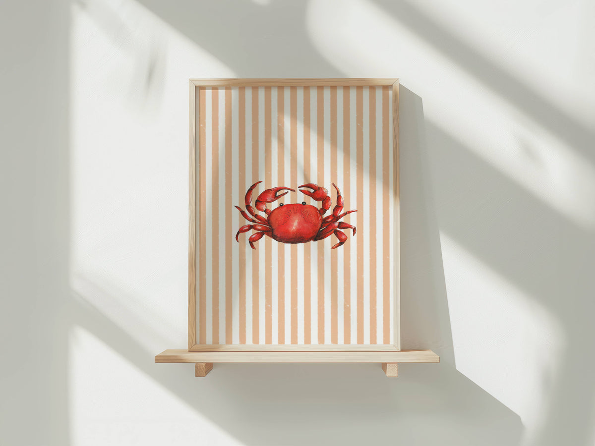 A framed art print featuring a red crab on a beige and white striped background, displayed on a light wood shelf against a white wall with soft window shadows.