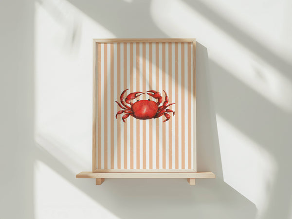 A framed art print featuring a red crab on a beige and white striped background, displayed on a light wood shelf against a white wall with soft window shadows.