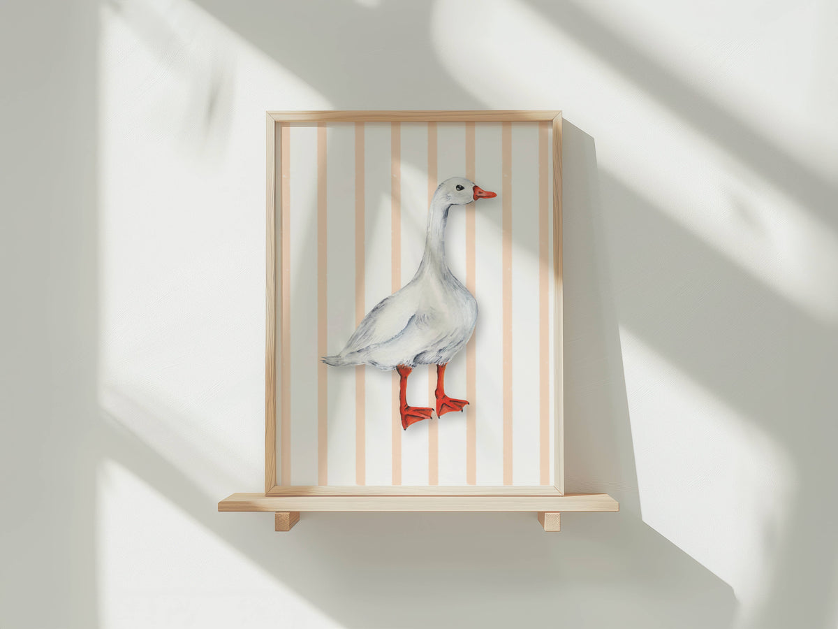A framed wall art piece displayed on a light wood shelf against a white wall, featuring a white goose standing in front of vertical peach and white stripes. The frame and shelf are light natural wood, and the goose illustration has a red beak and red legs.