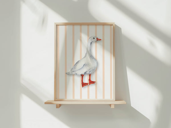 A framed wall art piece displayed on a light wood shelf against a white wall, featuring a white goose standing in front of vertical peach and white stripes. The frame and shelf are light natural wood, and the goose illustration has a red beak and red legs.
