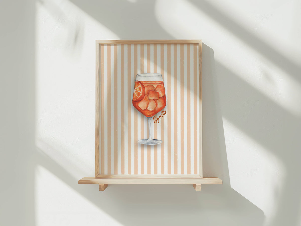 A framed art print featuring a watercolor illustration of a glass of orange spritz with citrus slices, displayed on a light wood shelf against a white wall with soft window shadows.
