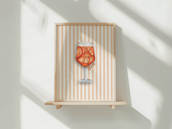 A framed art print featuring a watercolor illustration of a glass of orange spritz with citrus slices, displayed on a light wood shelf against a white wall with soft window shadows.