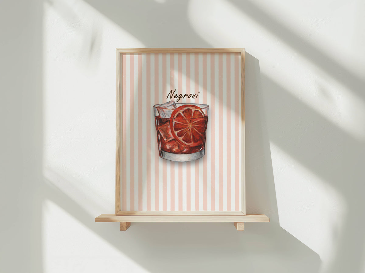 A framed art print featuring a watercolour illustration of a Negroni cocktail in a tumbler glass with ice and an orange slice garnish, set against a background of vertical coral and white stripes. The print is shown in a light wood frame displayed on a wooden shelf against a white wall with soft window shadows.